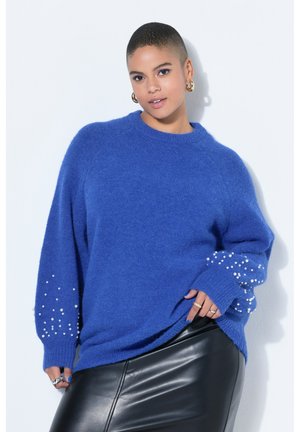 Kobieta o krótkich włosach, ubrana w niebieski sweter z perełkowymi zdobieniami na rękawach oraz czarną skórzaną spódnicę, pozująca na tle białej ściany.