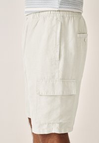 Light beige cargo shorts made of a textured fabric. Features include elastic waistband, side pockets, and a back button-closure pocket.