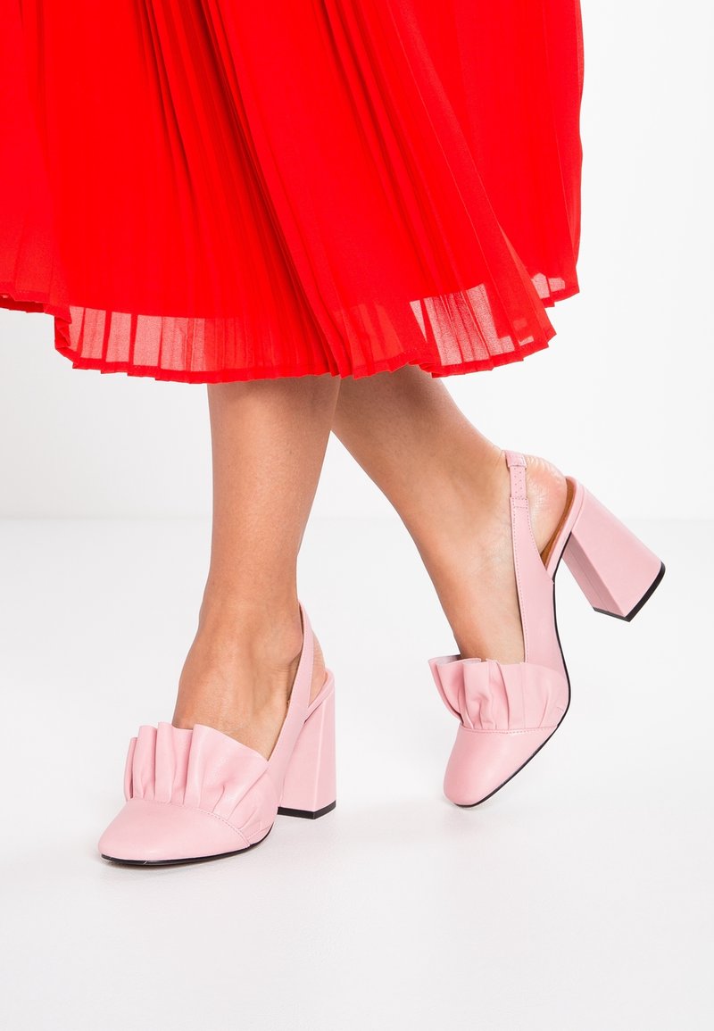 Femme portant une jupe plissée rouge et des chaussures slingback roses à talon bloc avec détail volanté, debout sur un sol blanc.