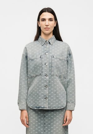 Kvinde iført et lyseblåt denim jakke- og nederdelssæt med subtilt gentagende hvidt monogrammønster, stående mod en ensfarvet hvid baggrund.