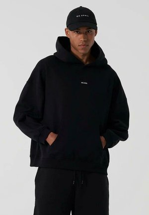 Black hoodie with a front pocket and a logo on the chest, paired with black pants and a matching cap. Soft, textured fabric.