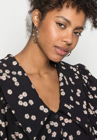Blusa preta com flores brancas e centros laranja, apresentando um decote em V, mangas bufantes e acabamento em renda. Brincos de argola prateados são visíveis.