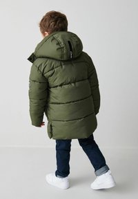 Cappotto verde oliva imbottito con un ampio cappuccio, colletto a coste e polsini elasticizzati. Presenta una toppa nera e uno spacco laterale per facilitare i movimenti.