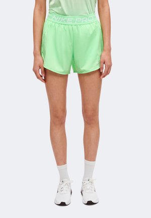 Žena v jasne zelených športových šortkách Nike Pro, bielych ponožkách a bielych bežeckých topánkach Nike, stojaca pred jednoliatym pozadím.