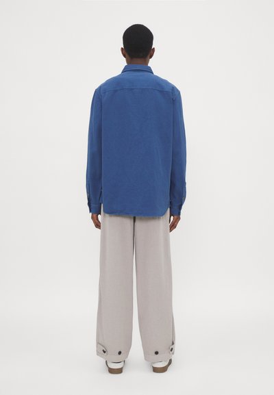 Camicione azzurro oversize con colletto, maniche lunghe e polsini con bottoni, abbinato a pantaloni larghi grigio chiaro con bottoni laterali.