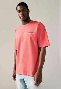 Koralfarvet bomulds t-shirt med korte ærmer, rund halsudskæring og trykte tekstdetaljer på brystet. Båret med lyseblå jeans.