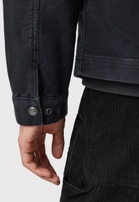 Manica di giacca blu scuro con bottone metallico, tessuto testurizzato e pantaloni in corduroy nero a coste visibili in basso. Mano appoggiata nelle vicinanze.