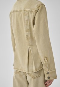 Beige Jeansjacke mit Rückenfalte und passende beige Jeanshose, getragen von einer Person vor einfarbigem Hintergrund.