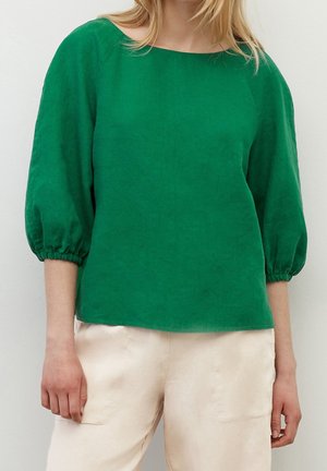 Vrouw draagt een groene blouse met lange mouwen en elastische manchetten en crème kleurige broek tegen een effen achtergrond.