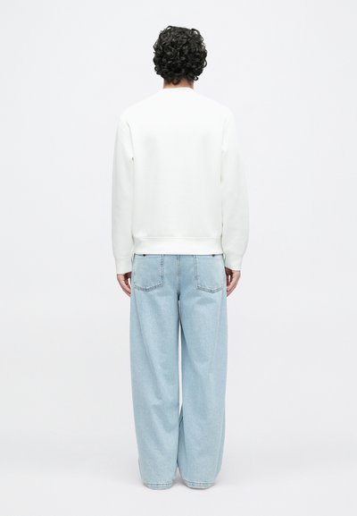 Λευκό φούτερ με στρογγυλή λαιμόκοψη και ριμπ μανσέτες, συνδυασμένο με ανοιχτό μπλε φαρδιά τζιν παντελόνια με πίσω τσέπες. Απλό φόντο.