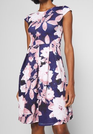 Femme portant une robe sans manches bleu marine avec un motif floral rose et blanc et une jupe plissée, debout devant un fond uni.