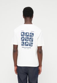 Άνδρας που φοράει λευκό μπλουζάκι με σχέδιο σε σκούρο μπλε αφηρημένο πλέγμα και κείμενο «Inhale Exhale Simple Pleasures» στην πλάτη, σε συνδυασμό με σκουρόχρωμο τζιν.