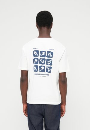 Mees kannab valget t-särki, millel on tagasi mereväesinise abstraktse ruudustiku muster ja tekst "Inhale Exhale Simple Pleasures", seljas on tumedad teksad.