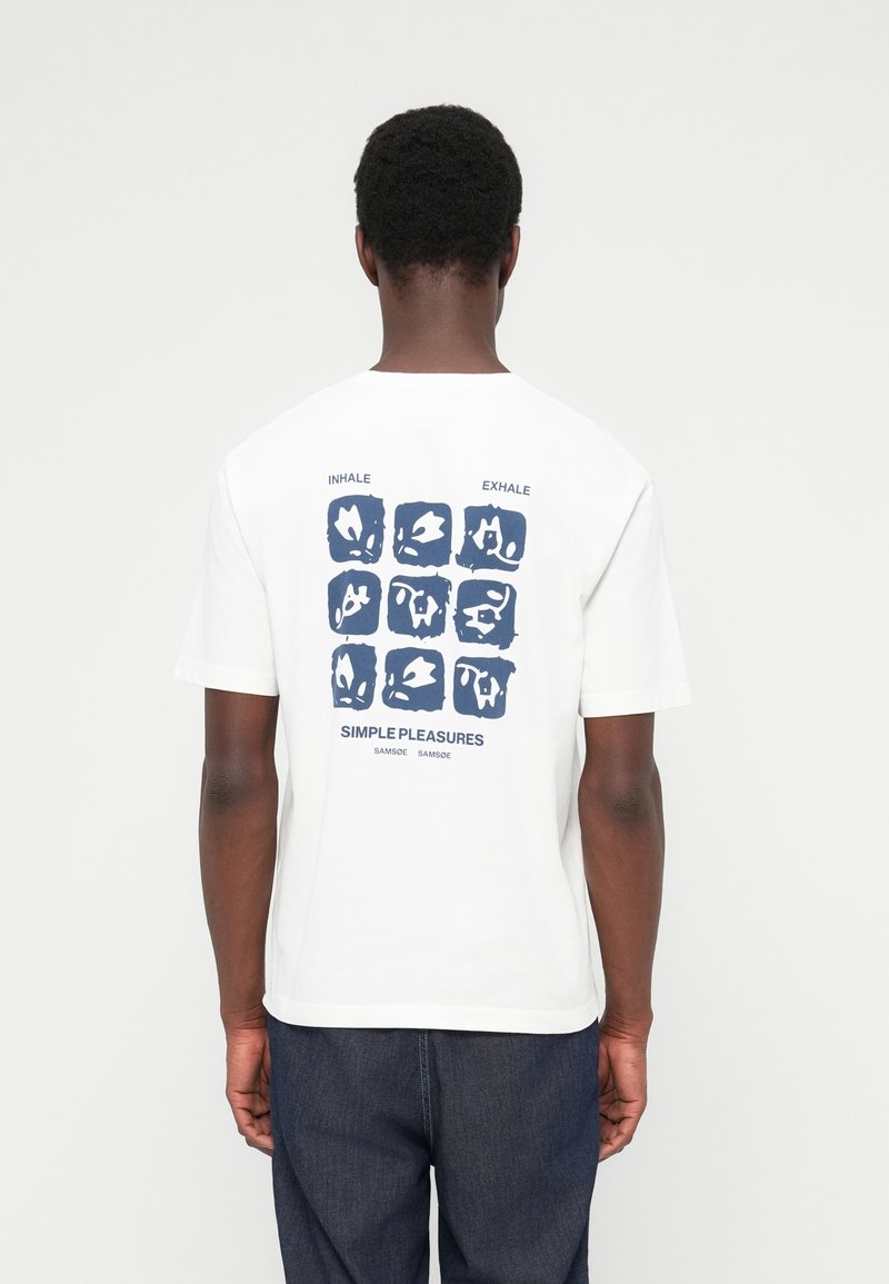 Άνδρας που φοράει λευκό μπλουζάκι με σχέδιο σε σκούρο μπλε αφηρημένο πλέγμα και κείμενο «Inhale Exhale Simple Pleasures» στην πλάτη, σε συνδυασμό με σκουρόχρωμο τζιν.