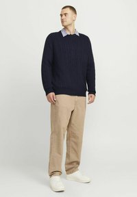 Tengerészkék cable-knit pulóver ribbelt mandzsettával, fekete világoskék ing fölött viselve, bézs nadrággal és fehér sportcipőkkel párosítva.