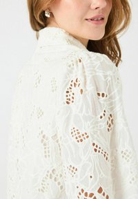 Hvit brodert bluse med intrikate blomsterdesign, gjennomsiktige områder og teksturert finish. Litt drapert design med myk krage.