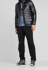 Sort pufferjakke med høj krave, grå grafisk t-shirt, sorte bukser og mørkegrønne vandresko med gule accenter.