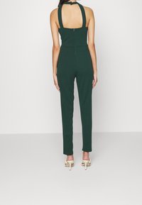 Mörkgrön jumpsuit med halterneck, öppen rygg, figursydd design och avsmalnande ben; har en bakre dragkedja och minimalistisk stil.