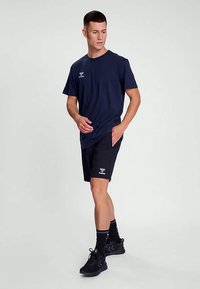 Sötétkék rövid ujjú póló és fekete rövidnadrág, mindkettő könnyű anyagból készült. Logó a pólón és a rövidnadrágon. A modellen fekete sportcipő van.