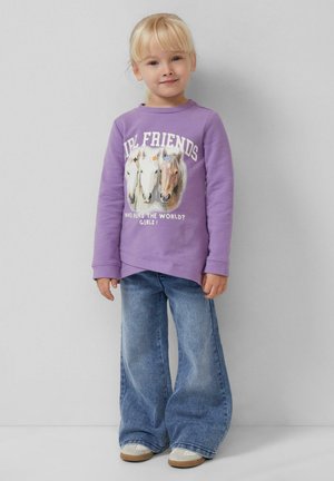 Junges Mädchen mit blonden Haaren, das einen lila Pullover mit Pferden und Text, weite blaue Jeans und weiße Turnschuhe trägt, steht vor einer grauen Wand.