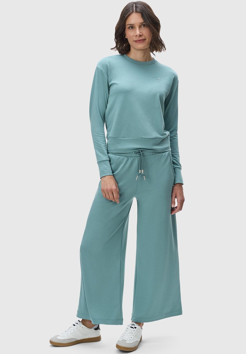 Completo a due pezzi in morbido tessuto teal. Comprende una felpa a maniche lunghe con colletto tondo e pantaloni larghi con vita elastica.