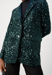 Smaragdgrön paljettblazer med tvåknappsstängning, slagkrage och framfickor, med en texturerad, glittrande yta.