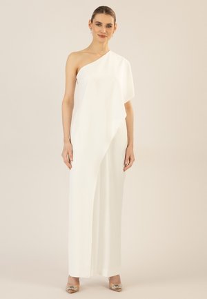 Woman in white one-shoulder jumpsuit with wide legs and embellished pointed heels, standing against a beige background.