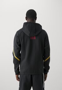 Schwarze Kapuzenjacke mit slim Fit, strukturiertem Stoff, roten und gelben Akzenten sowie drei vertikalen Streifen auf dem Rücken.