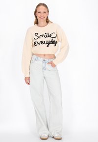 Przycięty beżowy sweter z czarnym napisem „Uśmiechaj się każdego dnia”, w połączeniu z jasnoniebieskimi spodniami w stylu wide-leg. Model nosi białe trampki. Gładka tekstura.