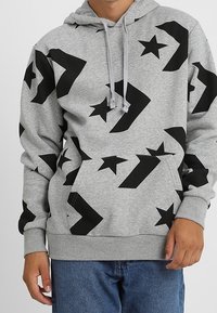 Gray hoodie featuring black geometric and star patterns, with a front pocket, drawstring hood, and ribbed cuffs and hem. Cotton blend material.