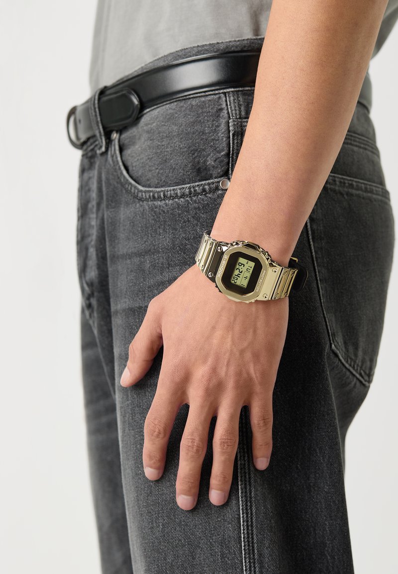 Digital klokke med rektangulært ansikt og digital skjerm, gullfarget metallarmbånd, brukt på en lys grå skjorte og mørk grå denimjeans.