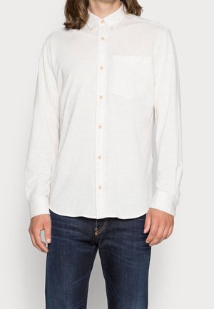 Mann trägt ein weißes, langärmliges Hemd mit Brusttasche und dunkelblaue Jeans, steht vor einem einfarbigen Hintergrund.