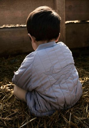 Barn med kort, mørkt hår iført en lys, quiltet jakke med fuglebroderi, sittende på halm vendt mot et gjerde i tre.
