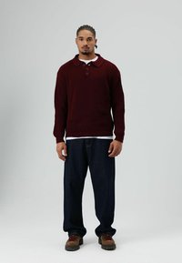 Jonge man met gevlochten haar die staat, gekleed in een bordeauxrode gebreide trui, donkere losse spijkerbroek, bruine wandelschoenen en een wit ondershirt.