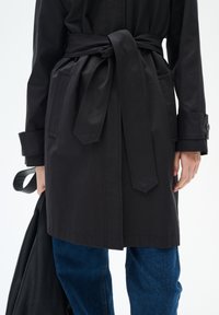 Svart trenchcoat med knytbart bälte, stora fickor och knäppta ärmar, kombinerad med blå denimbyxor. Slät tyg med skräddarsydd passform.