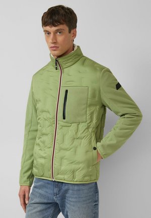 Man draagt lichtgroene gewatteerde jas met hoge kraag en borstzak, staand tegen een effen grijze achtergrond.