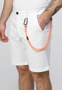 Pantaloni corti bianchi in cotone. Presentano una tasca frontale e una corda paracord arancione brillante con un moschettone nero attaccato in vita.