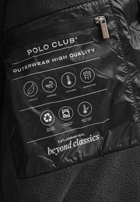 Svart tygetikett som visar skötselråd och funktionella egenskaper: ultralätt, vindtät, extra varm, vattenavvisande, tillverkad av återvunnen polyester.