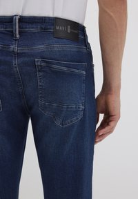 Jeans in denim blu caratterizzati da un lavaggio scuro, un classico design a cinque tasche e una patch in pelle nera nella parte posteriore della vita con il logo del marchio.