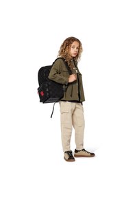 Zwarte rugzak met een monogram patroon, gedragen door een persoon in een olijfgroene shirt en beige cargobroek, met zwarte sneakers.