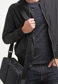 Svart bomberjacka med ribbade manchetter, en grå texturerad skjorta och en mörk budväska med silverdetaljer; denimjeans synliga.