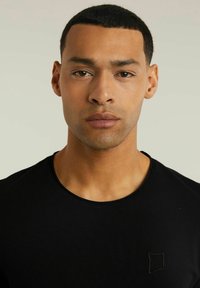 Fekete crew-neck póló, puha anyagból készült, bal oldalon egy kis hímzett négyzetlogóval. Simaság és testhezálló fazon jellemzi.
