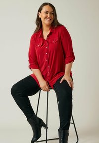 Rotes Button-up-Hemd mit zwei Brusttaschen, hochgekrempelten Ärmeln, getragen mit schwarzen Skinny Jeans und schwarzen Ankle Boots, sitzend auf einem Hocker.
