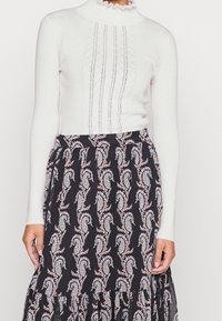 Weißer gerippter Rollkragenpullover mit fließendem Kragen, kombiniert mit einem schwarzen Rock mit bunten Paisleymustern und einem schichtweise angeordneten Saum.
