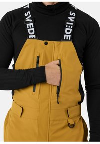 Gele waterdichte bretels met zwarte banden, voorritszakken en een verstelbare taille. Draag over een zwart aansluitend lange mouwen top.