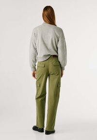 Vrouw met lang bruin haar draagt een lichtgrijs sweatshirt, olijfgroene cargobroek en zwarte schoenen, staand met de rug naar de camera op een effen achtergrond.