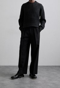 Pull en maille côtelée foncé avec col rond, associé à un pantalon noir ample et des chaussures noires vernies à lacets, debout devant un fond gris.