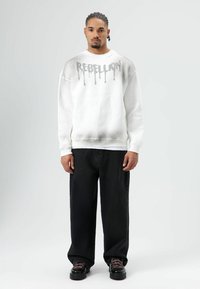 Witte sweatshirt met "REBELLION" in druipende grijze letters, gecombineerd met zwarte jeans met wijde pijpen en zwarte schoenen met roze accenten.