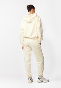 Kócos bunba kötött hajjal rendelkező nő krém színű kapucnis pulcsiban, passzoló melegítőnadrágban és fehér sportcipőben áll, keze a zsebében, háttal néz.