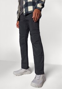 Pantalons zippés noirs en tissu léger et texturé, avec une longueur de jambe ajustable, associés à des chaussures de sport grises.